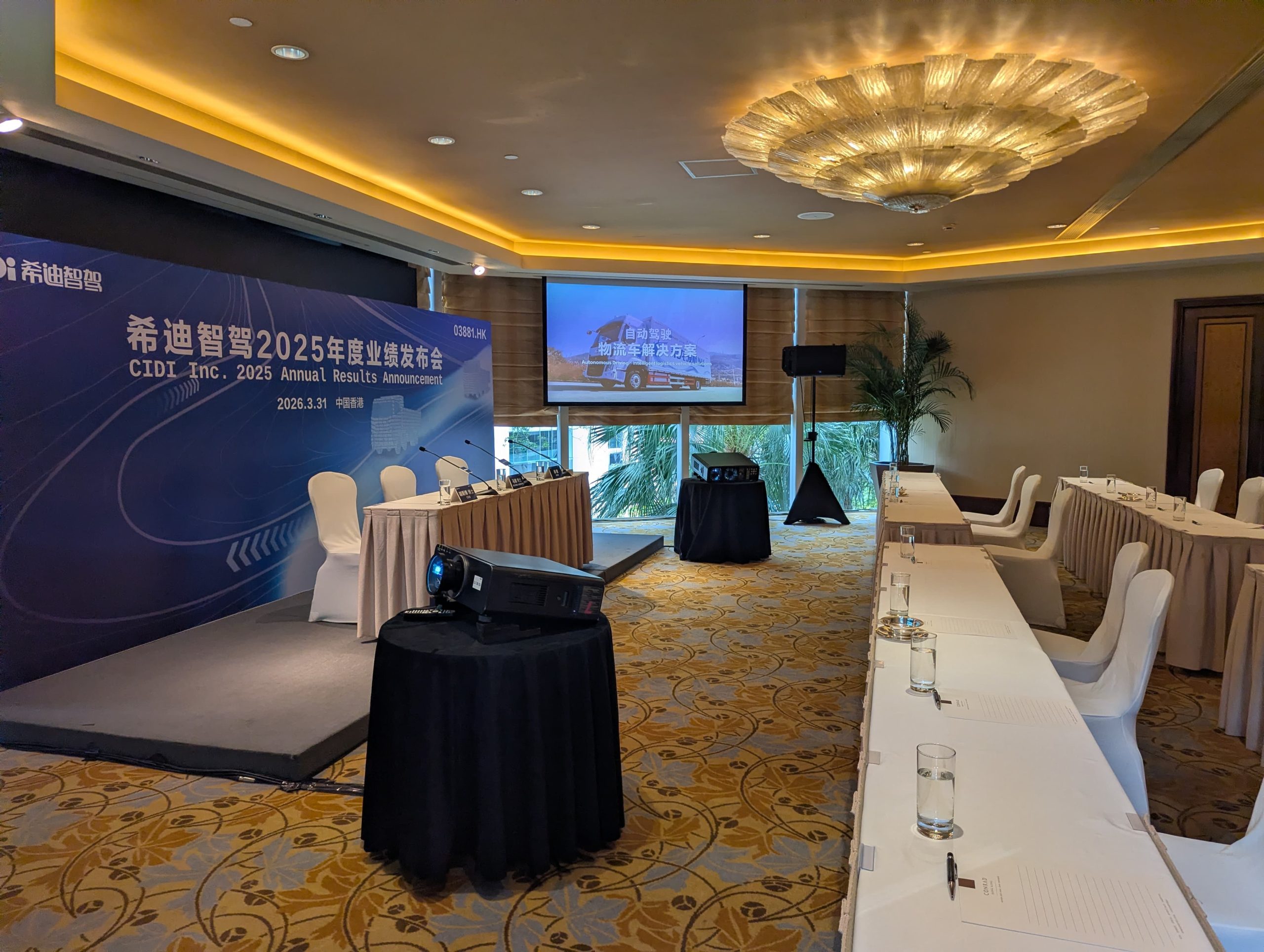 Small conference stage with a branded backdrop, projector, screen, speaker, and arranged meeting tables