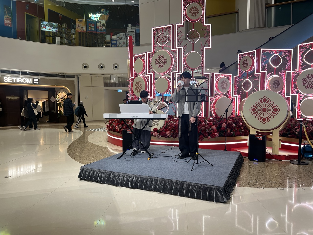 Musician performing on a small carpeted mall stage with a keyboard and microphone stands