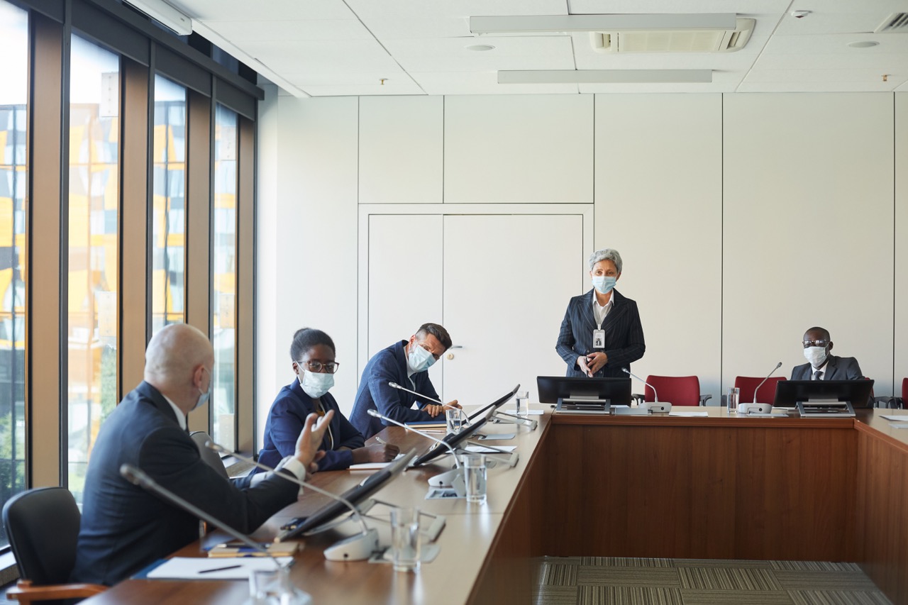 Masked participants in a formal conference room using tabletop microphones during a meeting