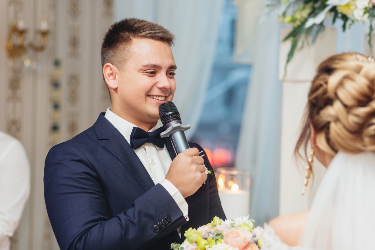 Man in a tuxedo speaking into a wireless microphone during a wedding reception