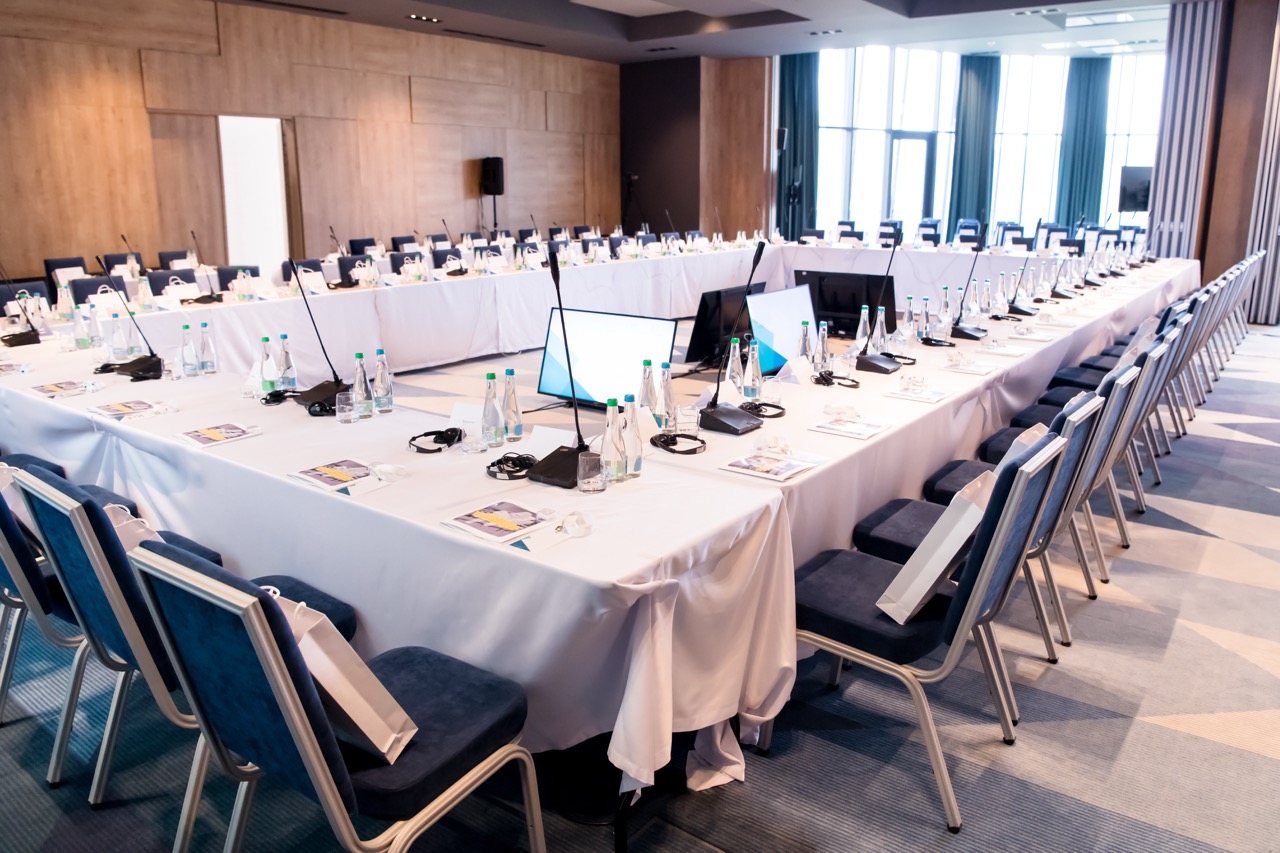 Empty conference room with U-shaped tables, tabletop microphones, monitors, and rows of chairs