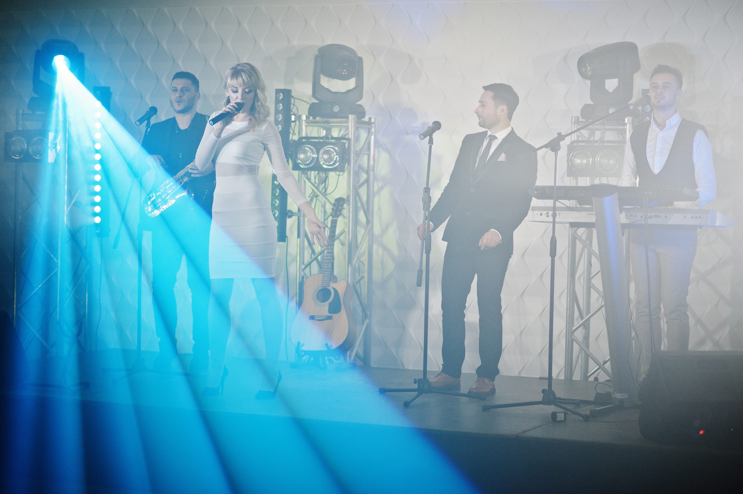Band performing on an indoor stage with microphones, keyboards and blue lighting